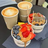 A high-angle, well-lit shot of two açaí bowls topped with fresh strawberries, blueberries, and peanut butter, accompanied by two lattes with latte art, served in branded paper cups on a black table.