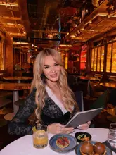 A portrait of a woman sitting at a table in a dimly lit, warm-toned bar or restaurant, featuring a cocktail and small plates of food in the foreground.