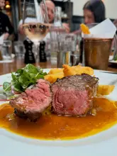 A candid, eye-level shot of a steak lunch at a restaurant, featuring a sliced steak with sauce and a side of fries, with a glass of wine visible in the background.