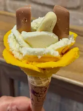 A close-up shot of an Amorino gelato rose cone, featuring mango and coconut flavors topped with chocolate pieces. The image captures the signature floral presentation of the brand against a neutral, blurred background.