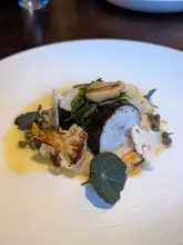 A high-quality, close-up photograph of a fine-dining fish dish, likely featuring white fish, mussels, and roasted cauliflower. The composition is clean, with a shallow depth of field that draws attention to the textures of the food. The lighting is professional, highlighting the plating details without harsh glare.