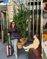 A lifestyle shot featuring a woman and a child with luggage outside the entrance of Hart Shoreditch, presented with a torn-paper collage effect that includes a glimpse of food on the right.