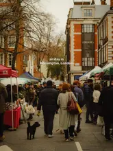 A candid, wide-angle street photography shot taken at a farmers market in Kensington, London. The image captures a crowd of people walking away from the camera on a grey, overcast day, with market stalls lining the street.