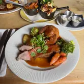 A high-angle, close-up shot of a Sunday roast dinner served on a white plate, featuring sliced roast beef, a large Yorkshire pudding, roasted potatoes, carrots, and broccoli, garnished with watercress. The setting appears to be a casual pub or restaurant, with additional side dishes visible in the background on a wooden table.