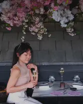 A polished, high-quality photograph featuring a woman dining at a restaurant decorated with cherry blossoms. The image captures a sophisticated, premium atmosphere, with the subject holding a glass of sparkling mocktail, perfectly aligning with the venue's 'Sakura' theme.