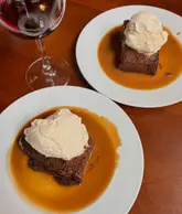 A top-down shot of two sticky toffee puddings served with ice cream and a glass of red wine on a wooden table.