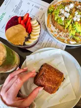 A bright, overhead flat-lay shot of a brunch spread at a cafe, featuring an acai bowl, avocado toast with feta, a pain au chocolat, and a matcha drink, captured in a clean, natural light setting.