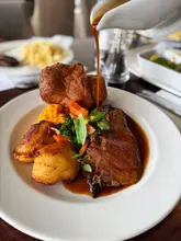 A high-quality, appetizing close-up shot of a Sunday roast dinner being served, featuring a dynamic gravy pour over the beef and vegetables. The lighting is natural and flattering, highlighting the textures of the food.