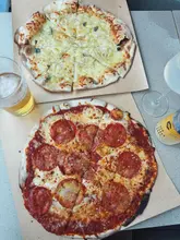 A top-down, flat-lay photograph of two pizzas—one spicy salami and one sage and truffle—served on brown paper alongside a glass of beer and a canned drink, set on a neutral table surface.
