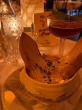 A close-up shot of a French onion soup served in a ceramic bowl, with a cocktail visible in the background. The lighting is dim and atmospheric, typical of a restaurant setting.