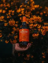 A high-quality, atmospheric product shot of a bottle of Deanston 17 Year Old whisky held in a hand, framed against vibrant orange flowers that match the label's colour palette.