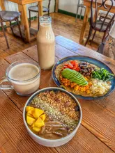 A high-angle shot of a plant-based brunch spread featuring a smoothie bowl, a tofu scramble dish, a latte, and a bottled smoothie on a wooden table. The image captures a healthy, wholesome meal in a cafe setting.