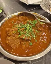 A close-up, top-down shot of a lamb curry served in a traditional metal balti dish, garnished with fresh coriander. The image focuses on the texture of the sauce and the meat, set against a dark table surface.