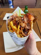 A close-up, handheld shot of a Greek gyro wrap, showcasing the meat and fries. The image is taken in a casual dining setting, likely near a window, providing natural lighting that highlights the texture of the food.