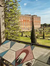 A bright, well-composed photograph taken at Hampton Court Palace, featuring an informative garden plaque in the foreground with the historic palace building and manicured gardens visible in the background.