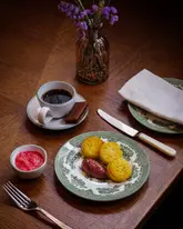 A beautifully composed, atmospheric shot of a refined breakfast or brunch service featuring crumpets with a fruit compote and a cup of black coffee, set against a dark, textured wooden table with elegant vintage tableware.