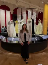 A clear, well-composed photograph of a creator standing in front of a display of royal dresses at the 'Queen Elizabeth II: Her Life in Style' exhibition at Buckingham Palace.