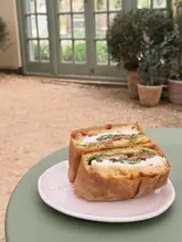 A close-up, eye-level shot of a fresh focaccia sandwich served in a rustic, garden-centre cafe setting. The sandwich is wrapped in brown paper and placed on a simple plate, with a soft-focus background of plants and a conservatory, creating an inviting and relaxed atmosphere.