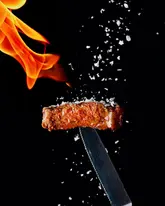 A high-end, dramatic studio photograph of a single slice of steak balanced on a knife, with flames in the background and salt falling over the meat. The image uses a stark black background to create a high-contrast, editorial look that emphasizes the texture and quality of the food.