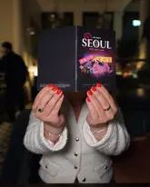 A person holds up a 'Seoul Food Tour Guide' menu from Six by Nico, obscuring their face. The image is taken in a dimly lit restaurant setting using a direct flash, which illuminates the menu clearly but leaves the background dark and creates a high-contrast look.