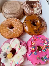 A top-down, flat-lay photograph of a box containing six assorted doughnuts, including glazed, chocolate-topped, and cinnamon-scroll varieties. The image captures the product in a casual, unstyled manner, likely intended to showcase a 'Too Good To Go' haul.
