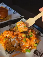 A close-up, high-angle shot focusing on a piece of food being lifted from a rice box with a wooden fork, with a burger visible in the blurred background.