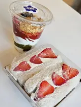 A top-down, 45-degree angle shot of a Japanese-style strawberry fruit sandwich and a granola yogurt pot, both still in their plastic takeaway containers, sitting on a plain white surface.