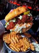 A close-up, high-contrast shot of a Sichuan-style fried chicken burger held in a hand, accompanied by a side of waffle fries. The image uses direct flash photography to highlight the textures of the food against a dark, blurred background.