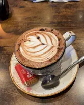 A clear, well-composed close-up shot of a cappuccino featuring latte art, served on a saucer with a Lotus Biscoff biscuit on a dark wooden table.