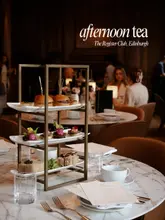 A sophisticated and well-composed photograph showcasing an afternoon tea service at The Register Club in Edinburgh. The image features a tiered stand filled with an assortment of savoury and sweet treats, set against a blurred, elegant restaurant interior, effectively communicating a premium dining experience.