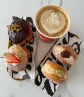 A high-quality, overhead flat-lay photograph of four artisan doughnuts and a latte, arranged on branded paper on a white marble surface. The lighting is bright and even, highlighting the textures of the doughnuts and the latte art.