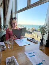 A woman sits at a table in the Cove Restaurant, holding a menu, with a bright, scenic view of the sea and coastline visible through the large window behind her.