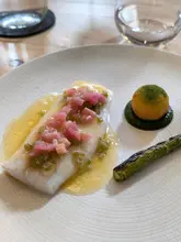 A high-quality, minimalist food photograph showcasing a fine dining dish of John Dory with rhubarb and asparagus. The lighting is soft and natural, highlighting the textures of the fish and the vibrant colours of the garnish. The composition is clean and professional, typical of high-end culinary photography.