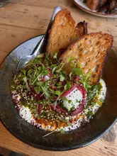 A high-quality, overhead shot of a spiced lamb and poached egg dish served with sourdough, featuring vibrant toppings like pickled onions and herbs.
