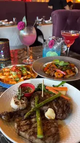 A montage video showcasing a variety of dishes and theatrical cocktails at a Pan-Asian restaurant. The video features close-up shots of food items like steak, noodles, bao buns, and spring rolls, alongside visually striking drinks with dry ice effects. The setting appears upscale with moody, ambient lighting.
