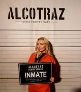 A promotional photo taken at Alcotraz, an immersive prison-themed cocktail experience. The subject is posing as an inmate against a mugshot backdrop, effectively showcasing the venue's unique branding and atmosphere.