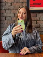 A high-quality, well-composed lifestyle portrait of a woman holding a refreshing cocktail in a restaurant setting. The image uses a shallow depth of field to keep the focus on the subject and the drink, with a clean, textured brick wall background that adds character without distracting from the main subject.