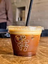 A high-quality, eye-level close-up shot of an iced coffee drink served in a clear plastic cup with a branded logo. The drink features a distinct, thick layer of whipped espresso foam resting on top of the iced coffee and milk, set against a wooden table surface.