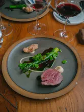 A high-quality, top-down shot of a fine dining main course featuring rose veal, sweetbreads, asparagus, and broad beans, served on a rustic ceramic plate. The composition is clean and professional, with wine glasses in the background adding to the dining atmosphere.