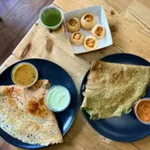 A top-down, overhead shot of a casual Indian street food spread featuring dosas, pani puri, and various chutneys served in takeaway-style containers on a wooden table.