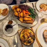 An overhead shot of a Sunday roast spread, featuring a large platter of roast potatoes, red cabbage, carrots, and parsnips, alongside a cast-iron skillet of cheesy leeks and broccoli, and individual plates with pork belly.