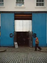 A moody, atmospheric, and well-composed shot of a person walking past a large, industrial blue warehouse door with 'G' and 'B' painted on it. The scene includes an 'Open' sign indicating the venue serves coffee, food, and wine, creating an inviting and aesthetic vibe.
