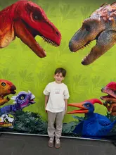 A photo of a young child standing in front of a branded dinosaur-themed backdrop, likely at an event or activity venue. The image is a straightforward, casual portrait capturing a family day out.