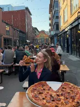 A lifestyle shot capturing a woman enjoying a slice of pizza at an outdoor venue, likely following a marathon event. The image uses natural golden hour lighting to create an inviting, social atmosphere.