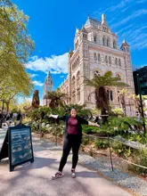 A bright, clear, and well-composed photograph of a visitor standing in front of the iconic Natural History Museum in London. The image captures the grandeur of the architecture and the surrounding gardens, effectively showcasing the location as a popular tourist destination.