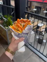 A handheld, close-up shot of a Greek gyro wrap held in front of an outdoor seating area at St James Stack. The focus is on the crispy fries and sauce topping the wrap, presented in branded paper.