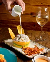 A high-quality, professional food photography shot capturing the dynamic action of pouring a sauce over a beautifully plated Peruvian ceviche dish, accompanied by a glass of white wine. The lighting is soft and intentional, highlighting the textures of the ingredients.