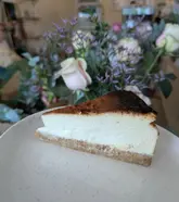 A high-quality, close-up photograph of a slice of Basque cheesecake on a speckled plate, featuring a soft-focus floral background that creates an inviting, cafe-style atmosphere.
