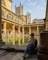 A high-quality, well-composed travel photograph featuring a visitor sitting on the edge of the Roman Baths in Bath, UK. The image captures the historic architecture and the iconic green water of the baths, with the subject positioned to draw the viewer's eye into the scene.