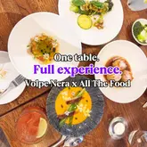 A high-quality, overhead flat-lay photograph showcasing a multi-course dining experience at Volpe Nera. The image features several refined, artistically plated dishes and a cocktail, arranged neatly on a wooden table, highlighting the restaurant's sophisticated culinary presentation.